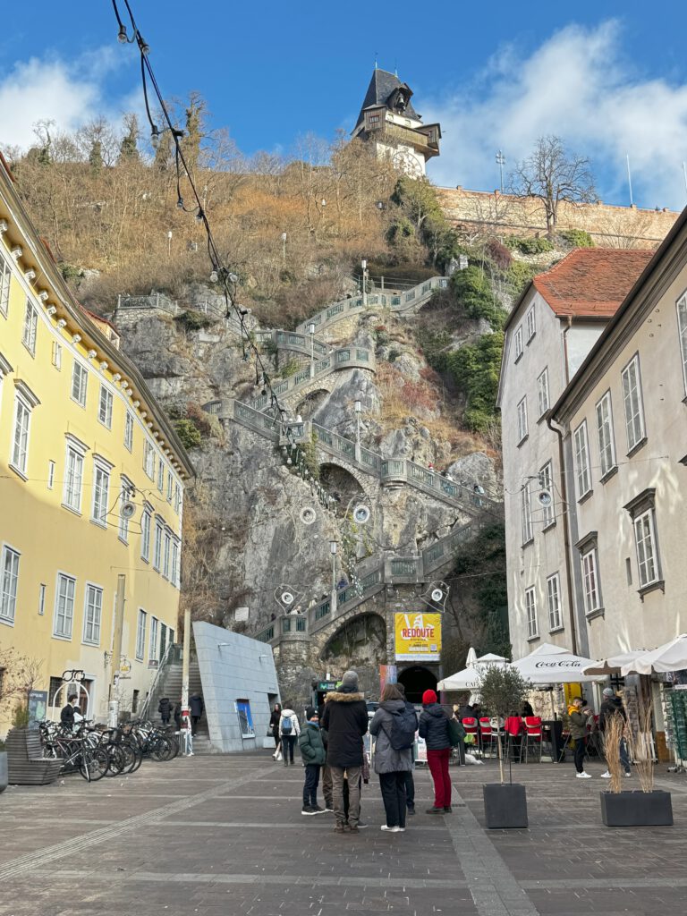 Blick von unten auf den Schlossberg. Es führt eine Steile Treppe im Fels nach oben. Einige Leute stehen unten und schauen nach oben. Man sieht den Grazer Glockenturm von unten.