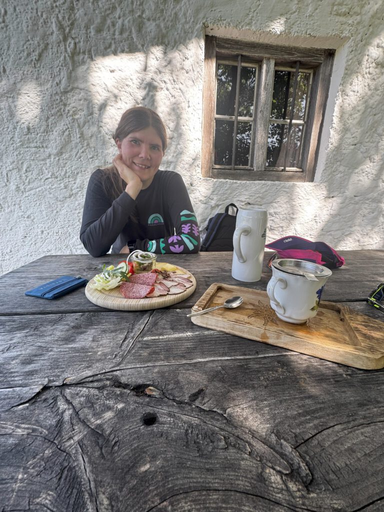 Eine Frau sitzt bei der Bäckeralm an einem Tisch. Auf dem Tisch steht ein Tee, ein Krug Buttermilch und ein Brotzeitbrettl mit Wurst und Käse.