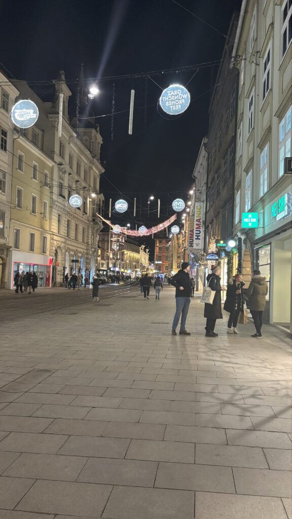 Zu sehen ist Graz bei Nacht. Die Straße ist festlich beleuchtet. Es sind sind wenige Menschen auf der Straße.
