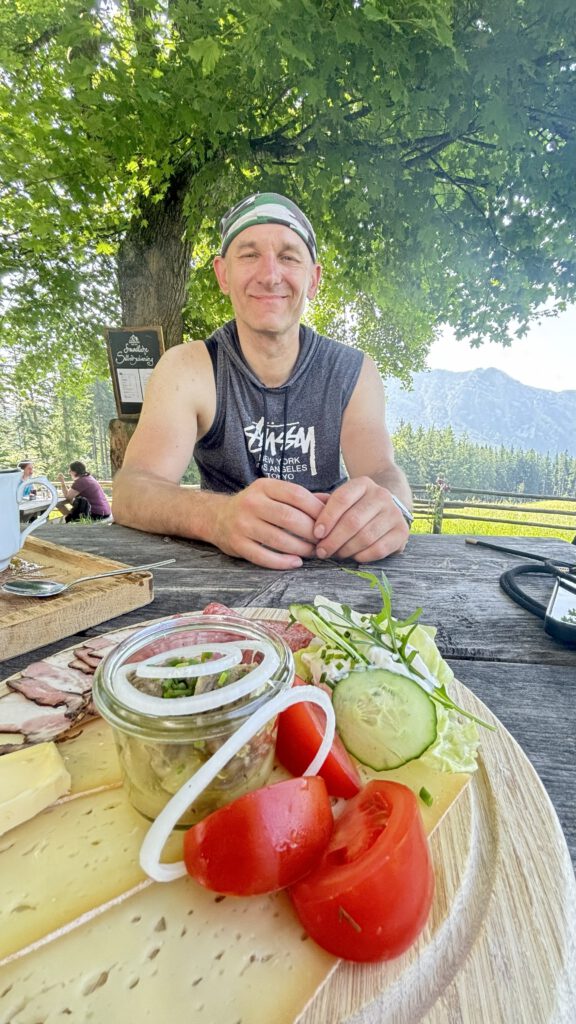 Es ist ein Mann zu sehen der an einem Tisch bei der Bäckeralm sitzt. Vor ihm ein Brotzeitbrett mit Käse, Wurst, Tomate und Gurke.